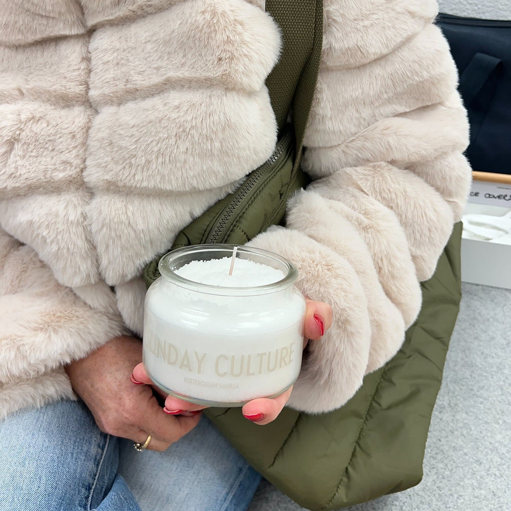 Person holding a candle labeled 'Sunday Culture' wearing a fluffy white jacket.