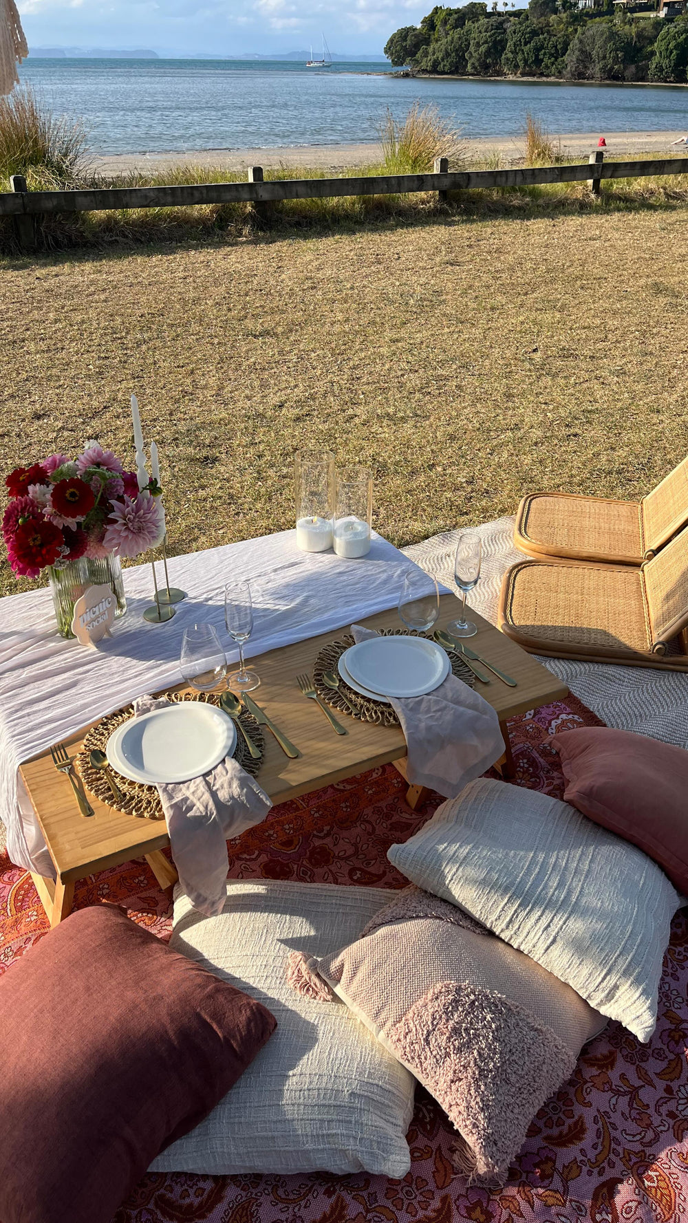 Outdoor picnic setup with table, plates, and pillows by a waterfront.