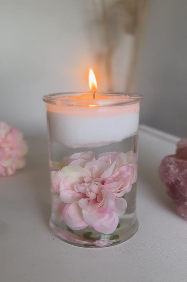 Candle in a glass holder with pink flowers floating on water on a white surface.