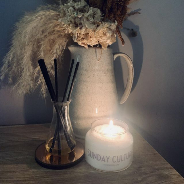 Decorative items including a vase with dried flowers, diffuser, and a sand wax sunday culture candle on a wooden surface.